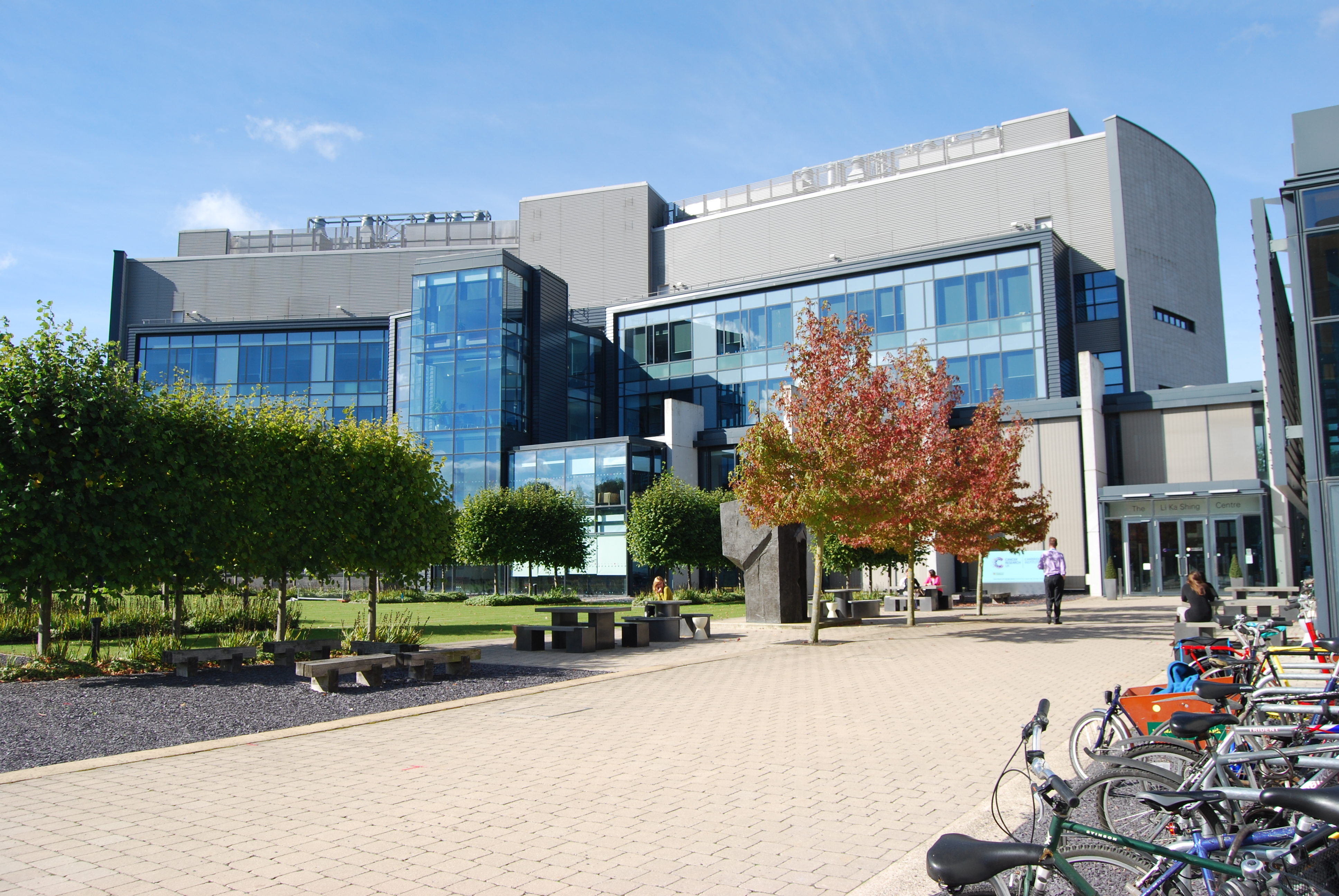 Cancer Research UK Cambridge Institute, a glass-fronted grey building in the daytime, with a row of trees to the left and cycle racks with bikes to the right of the pathway.