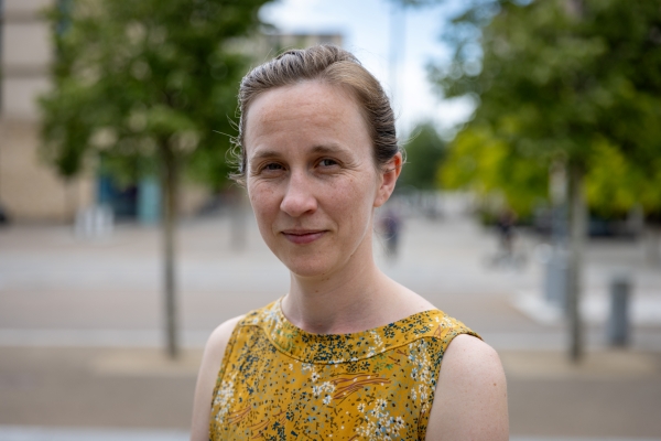 Headshot of Aspen Reese, a smiling blonde woman in a yellow top, standing outside in front of two trees.