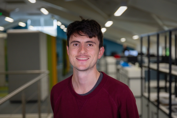 Headshot of Charlie-Barty King, a smiling brunette man in a red jumper.