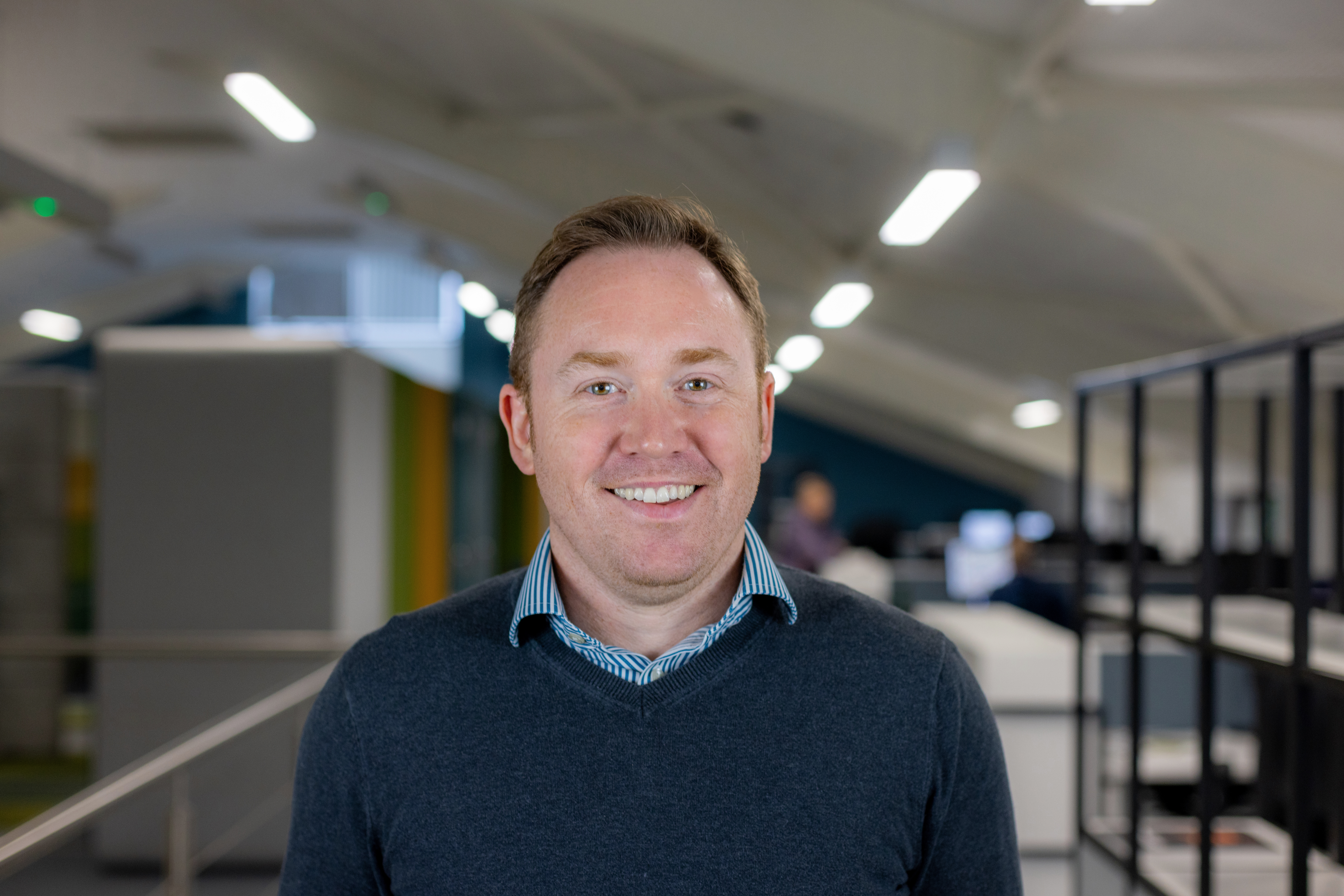 Headshot of Greg Nolan, a smiling man in a navy jumper and blue shirt.