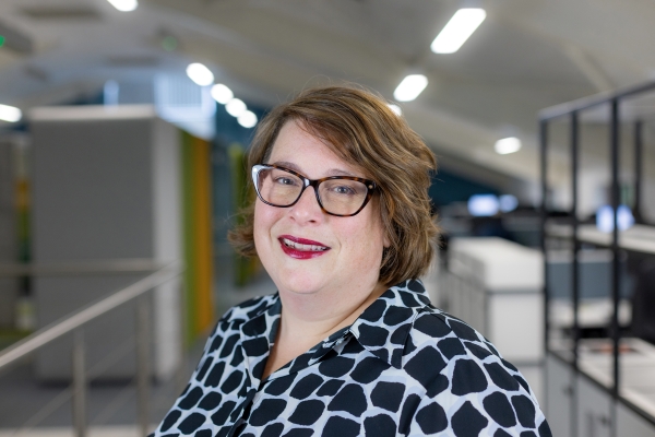 Headshot of Jo Beale, a smiling brunette woman with glasses in a black and white shirt.