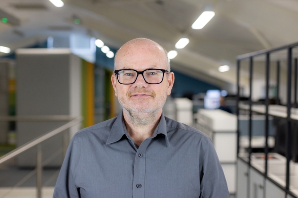 Headshot of Max Shone, a smiling man with black glasses in a grey shirt.